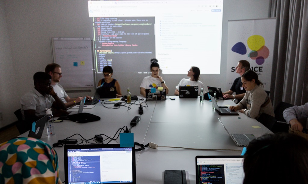 Teilnehmende beim Python-Workshop mit Laptops am Tisch, im Hintergrund Leinwand mit Code.